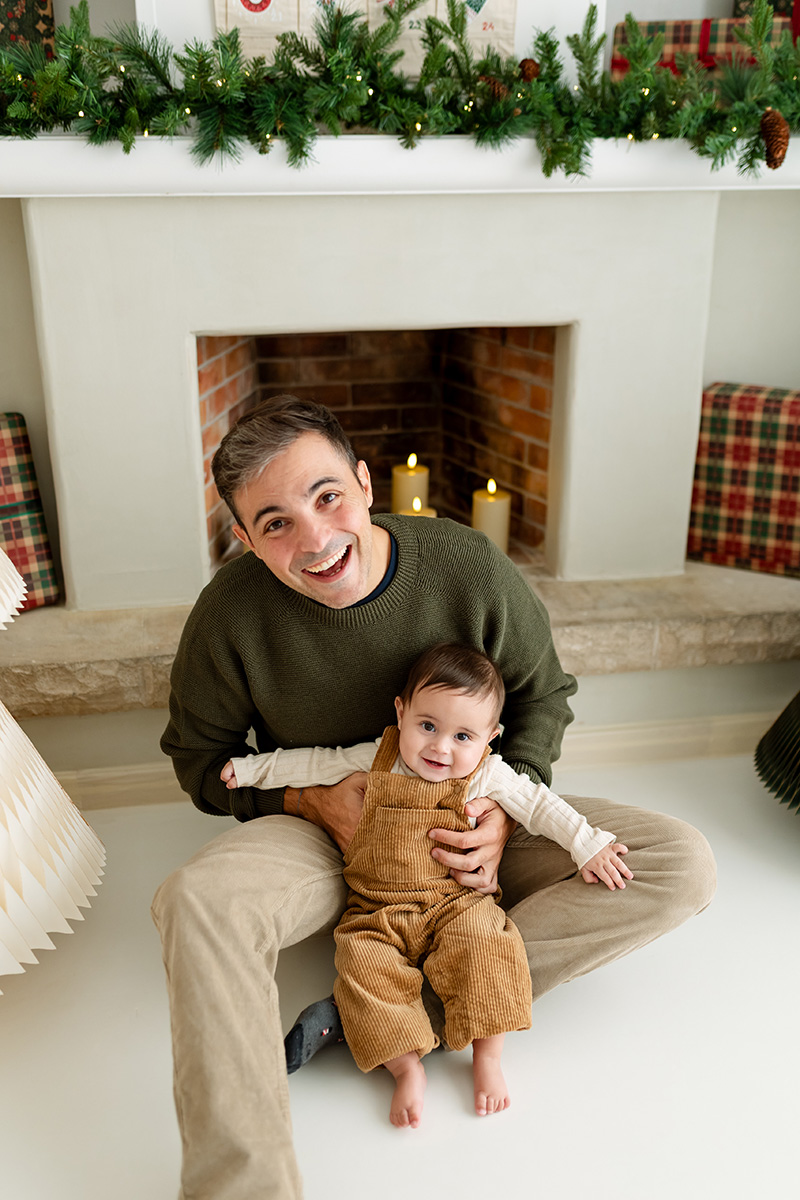 Papá sentado en el suelo con su bebé sonriendo a cámara delante de chimenea Navidad