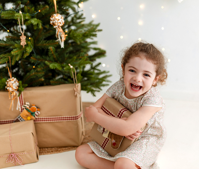 niña riéndose cogiendo un regalo con un árbol de navidad y luces de fondo