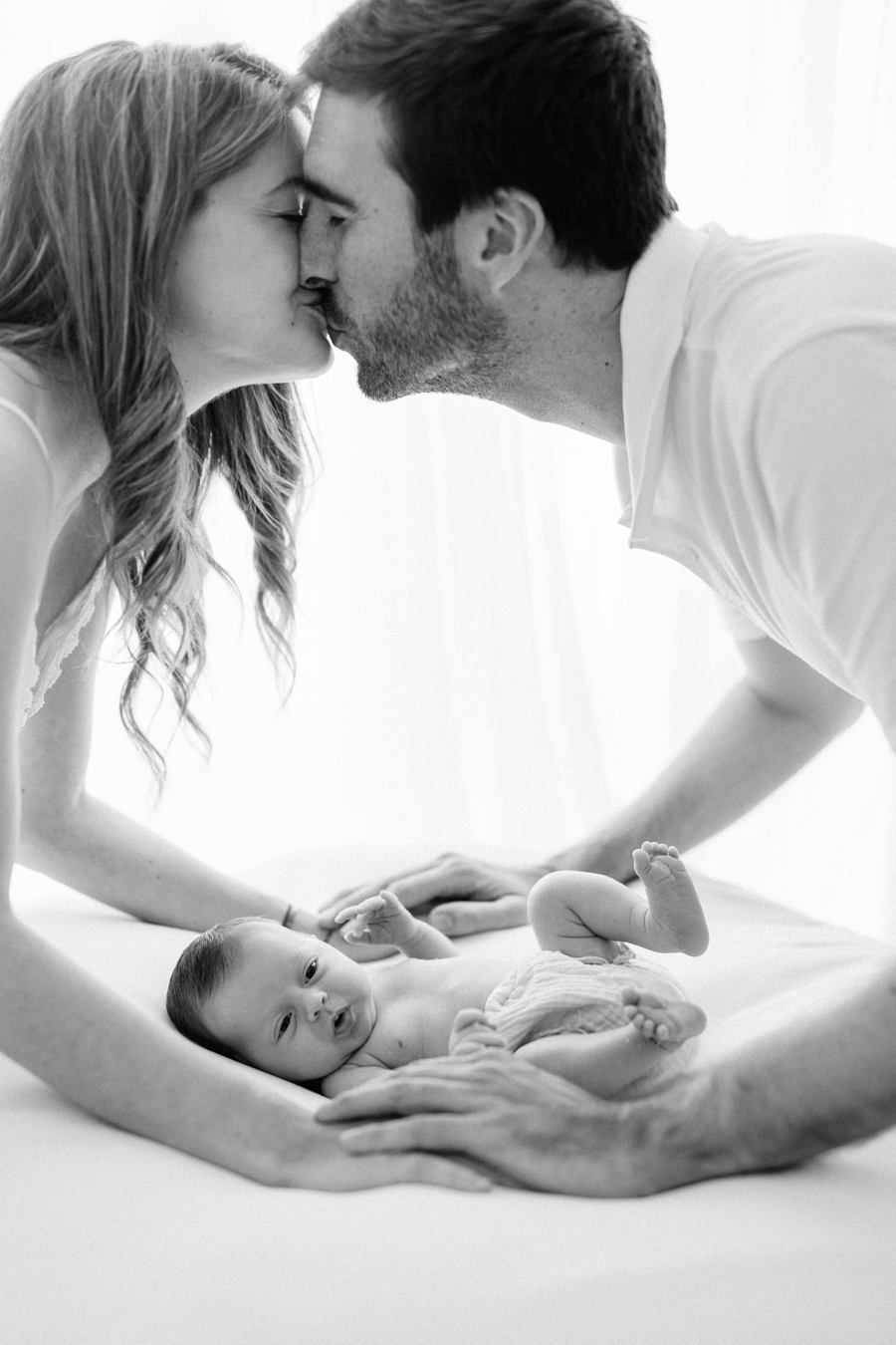 Padres besándose mientras su bebé recién nacido descansa en la cama durante una sesión de fotos newborn natural en familia en Barcelona.