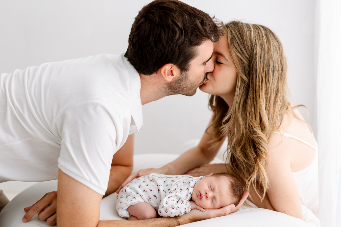 Retrato familiar en Barcelona con padres besándose mientras sostienen a su bebé recién nacido en una sesión de fotos newborn natural.