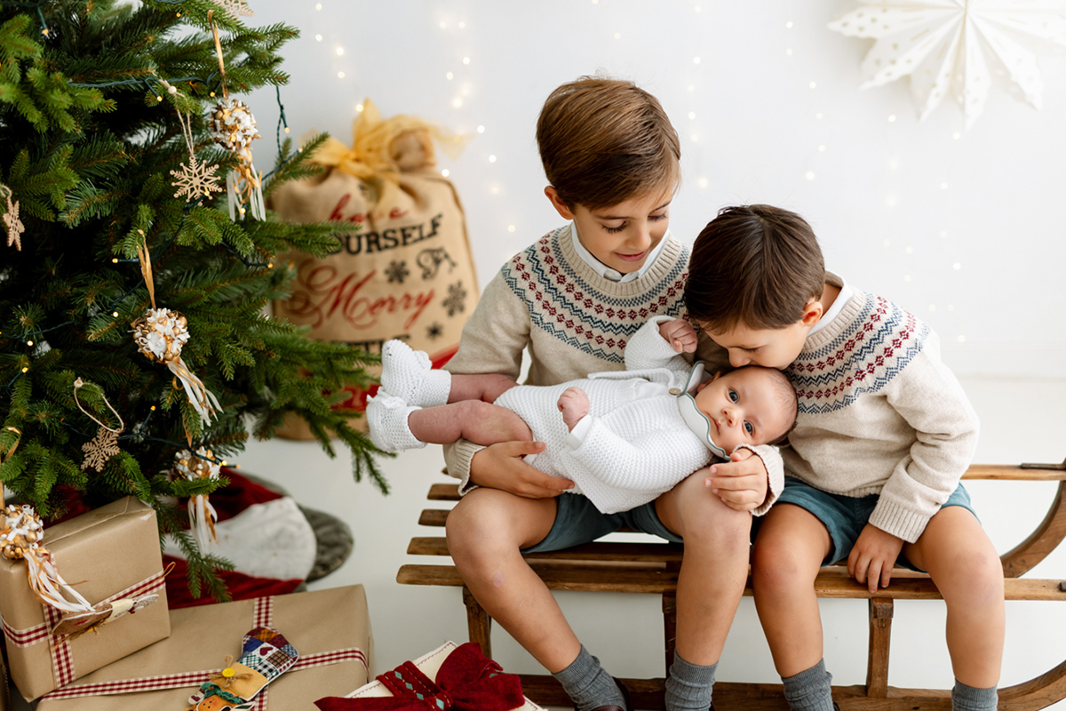 Hermanos mayores besando y cogiendo a su hermano recién nacido en sesión de fotos de Navidad en Barcelona